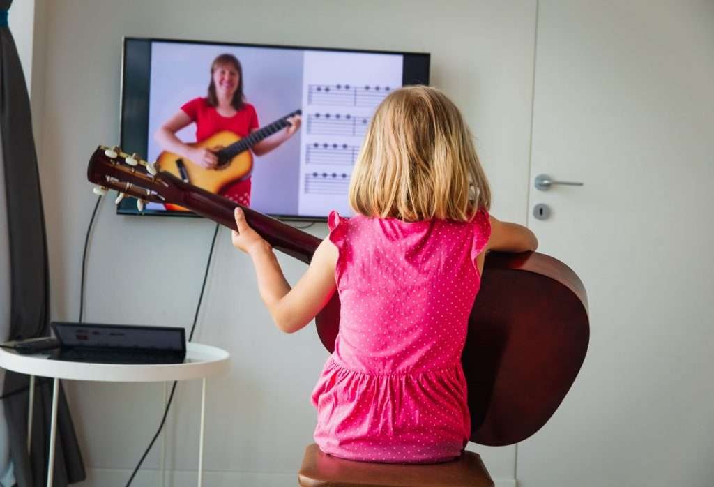 little girl playing guitar while watching her teacher play guitar on a big screen tv